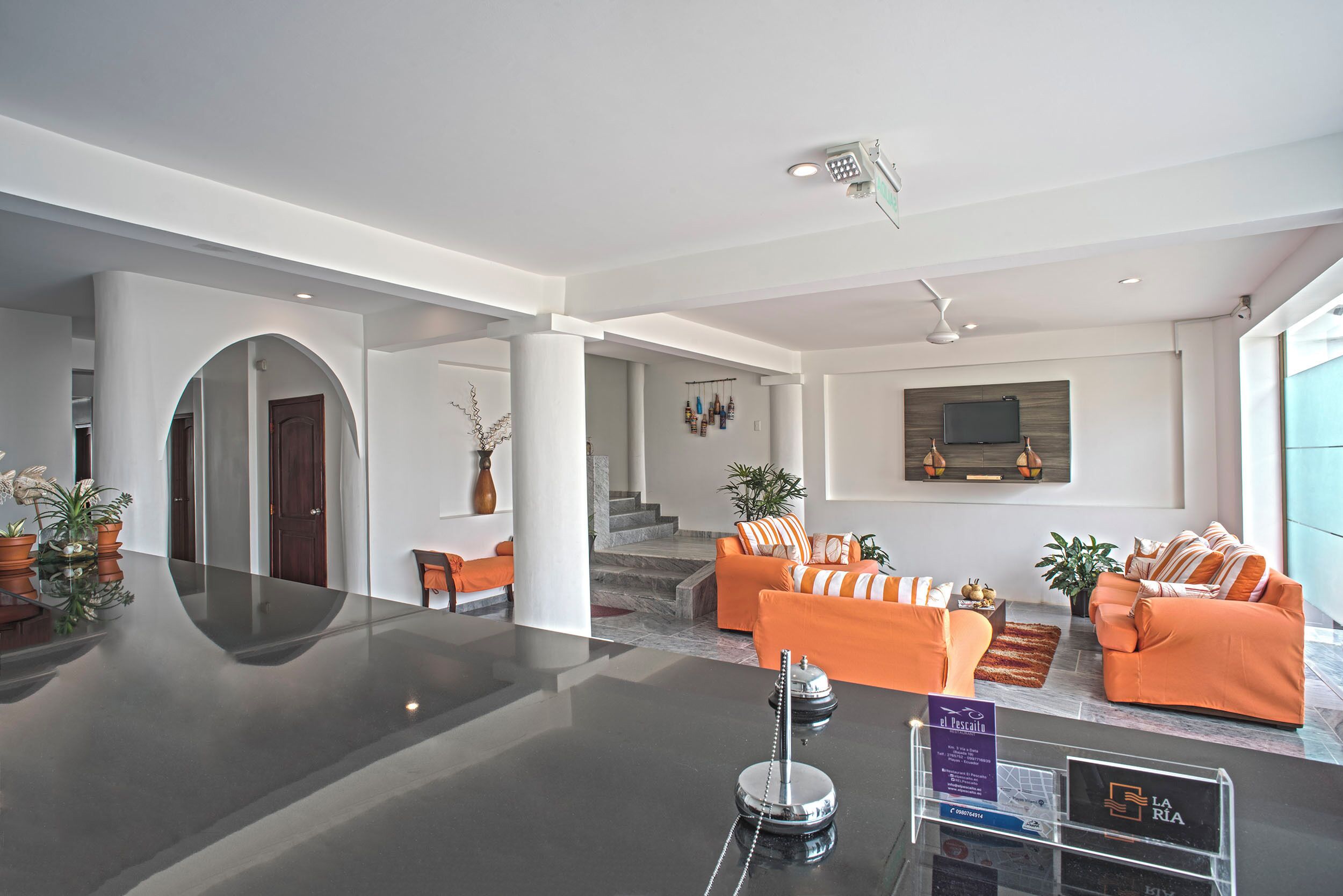 Un vestíbulo amplio con diseño moderno, que incluye una zona de estar central con sofás naranja, un mostrador de recepción y una escalera que conduce a los pisos superiores.