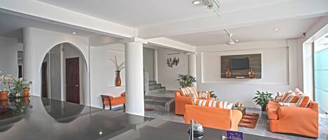 A spacious lobby with a modern design, featuring a central seating area with orange sofas, a reception desk, and a staircase leading to upper floors.