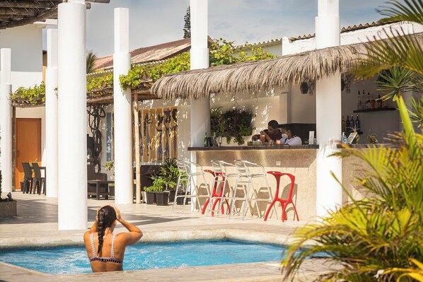 Zona de piscina con bar y asientos, rodeada de pilares blancos y un techo de paja.