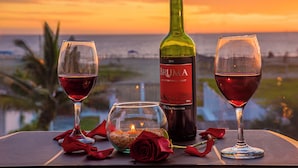 Taula amb dues copes de vi, una ampolla de vi Bruma, una espelma i una rosa a un balcó amb vista al mar al capvespre.