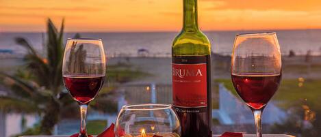 Une table avec deux verres à vin, une bouteille de vin Bruma, une bougie et une rose, sur un balcon donnant sur l’océan au coucher du soleil.