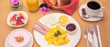 Desayuno con huevos revueltos, jamón, queso, tostadas, fruta, yogur y café.