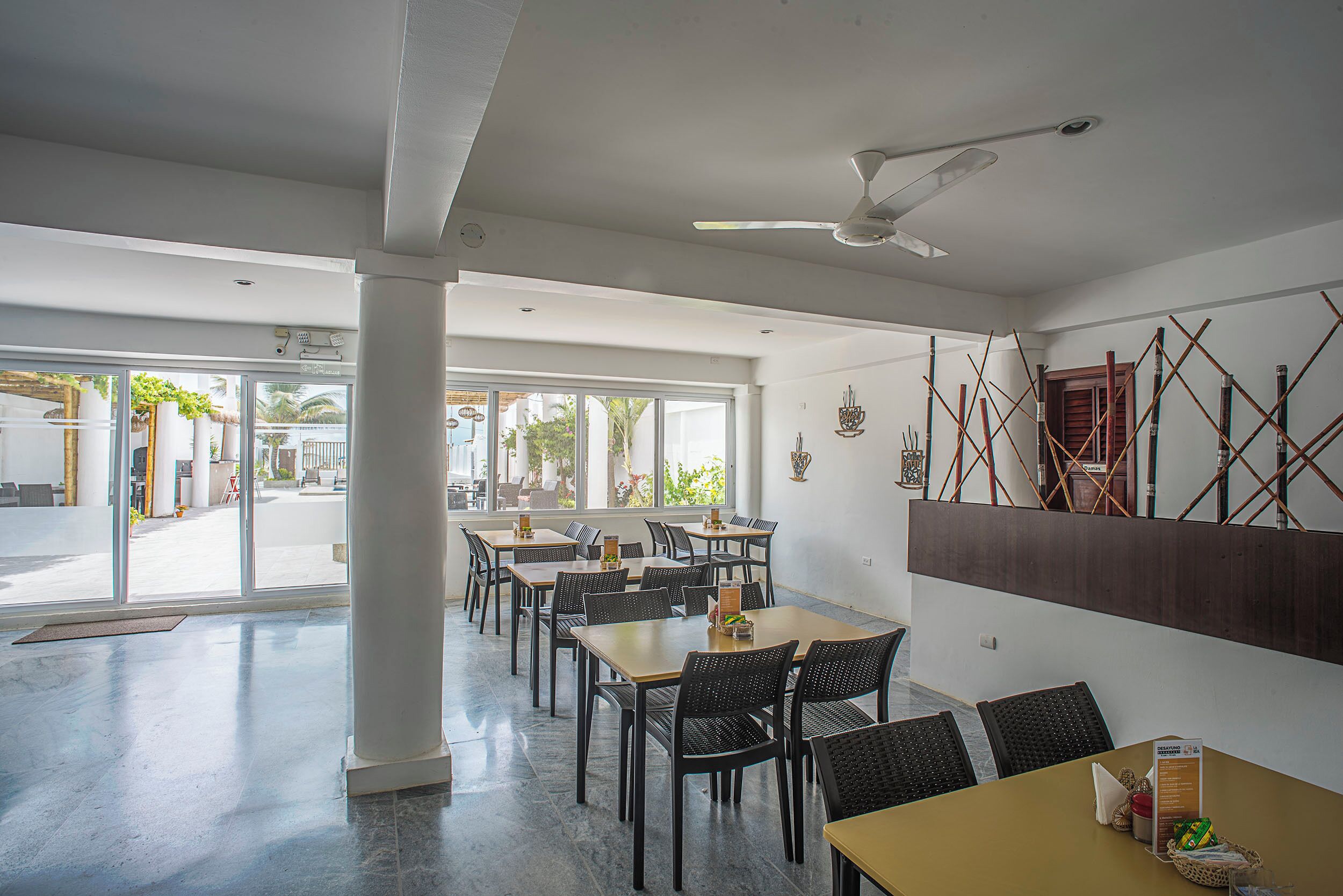 A spacious dining area with high ceilings, large windows, and a ceiling fan. Tables and chairs are arranged for dining.