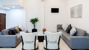 A modern living room with two grey sofas, white chairs, a small table with a plant, and a TV mounted on the wall.