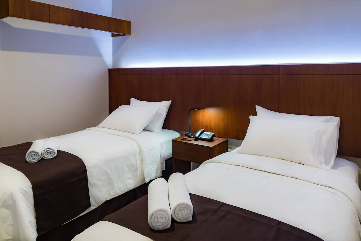A hotel room with two beds, each with white linens and brown headboards. There is a nightstand with a lamp and a phone between the beds.