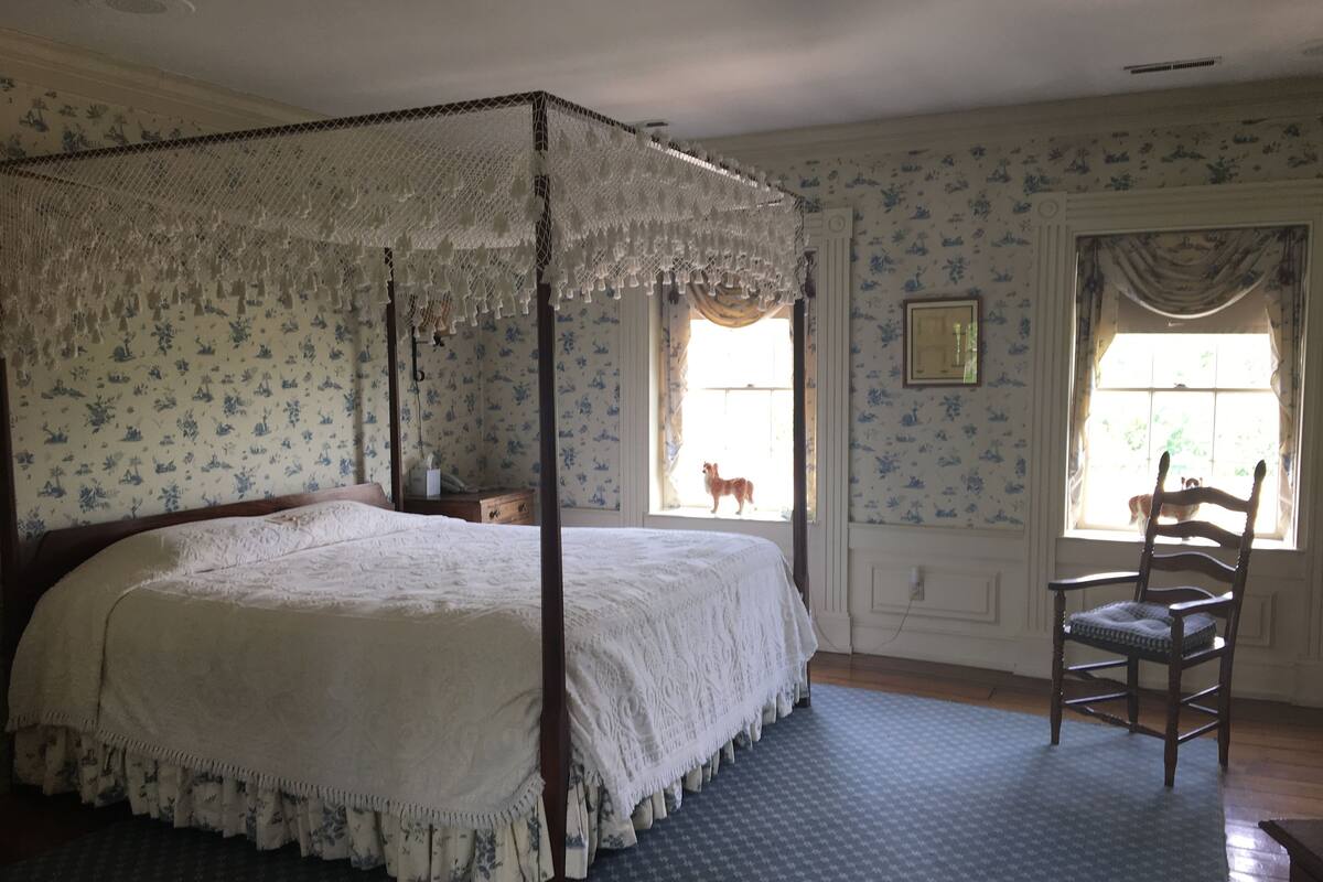 Un lit à baldaquin à quatre poteaux, surmonté d’un dais en dentelle, une chaise en bois et un tapis à motifs.