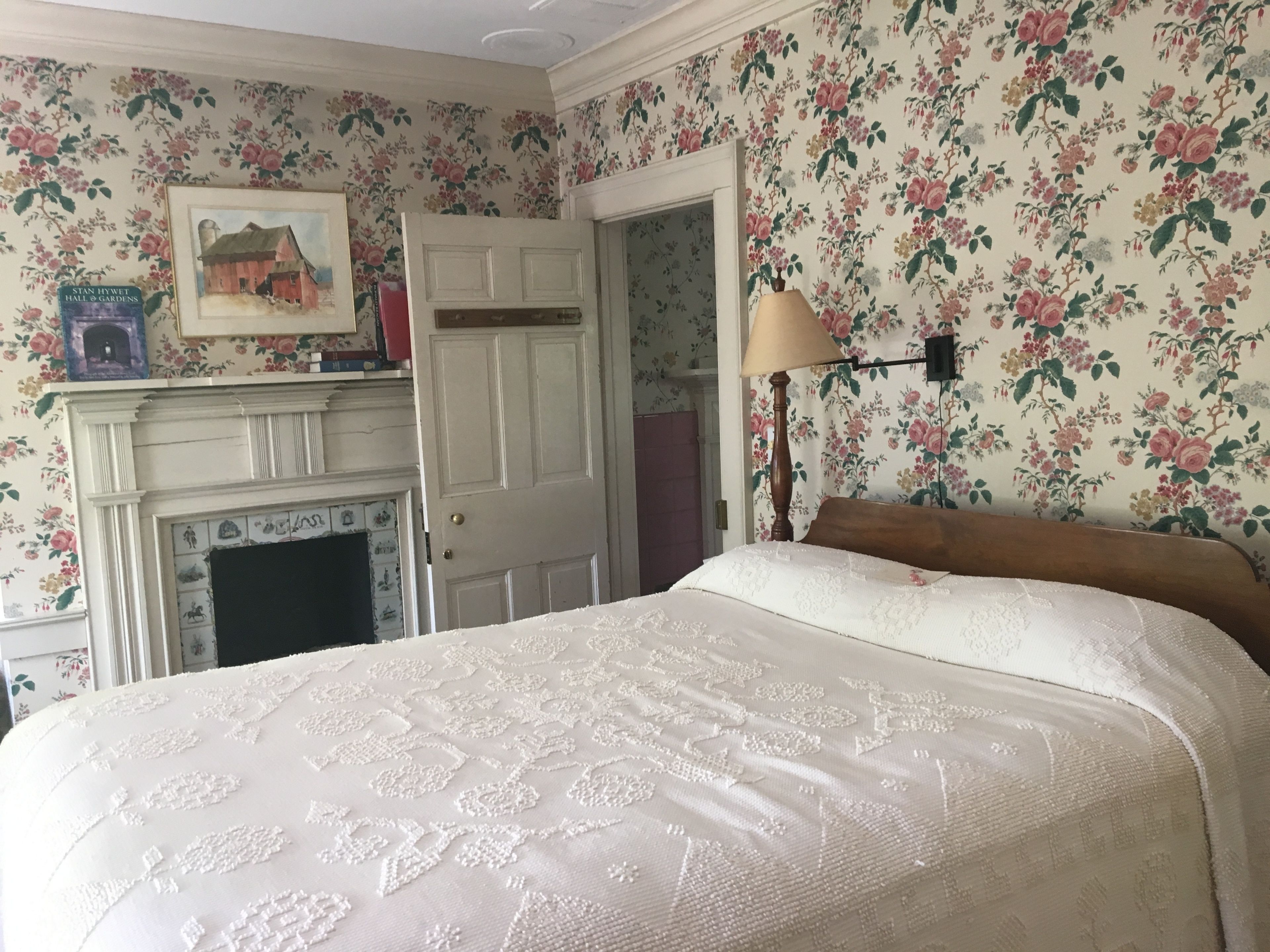 Un dormitorio con papel tapiz floral, una cama con cubrecama blanco bordado, una mesita de noche con lámpara y una chimenea con repisa.