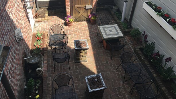 Un patio con suelo de ladrillo, sillas de metal, una mesa, plantas en macetas y una puerta de madera.