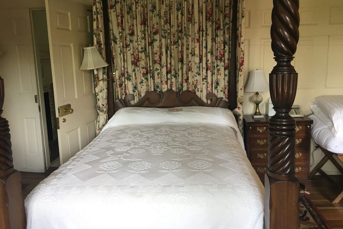 Un lit traditionnel à baldaquin, agrémenté d’un rideau à motifs floraux, une table de chevet avec une lampe et une petite table de nuit avec un téléphone.