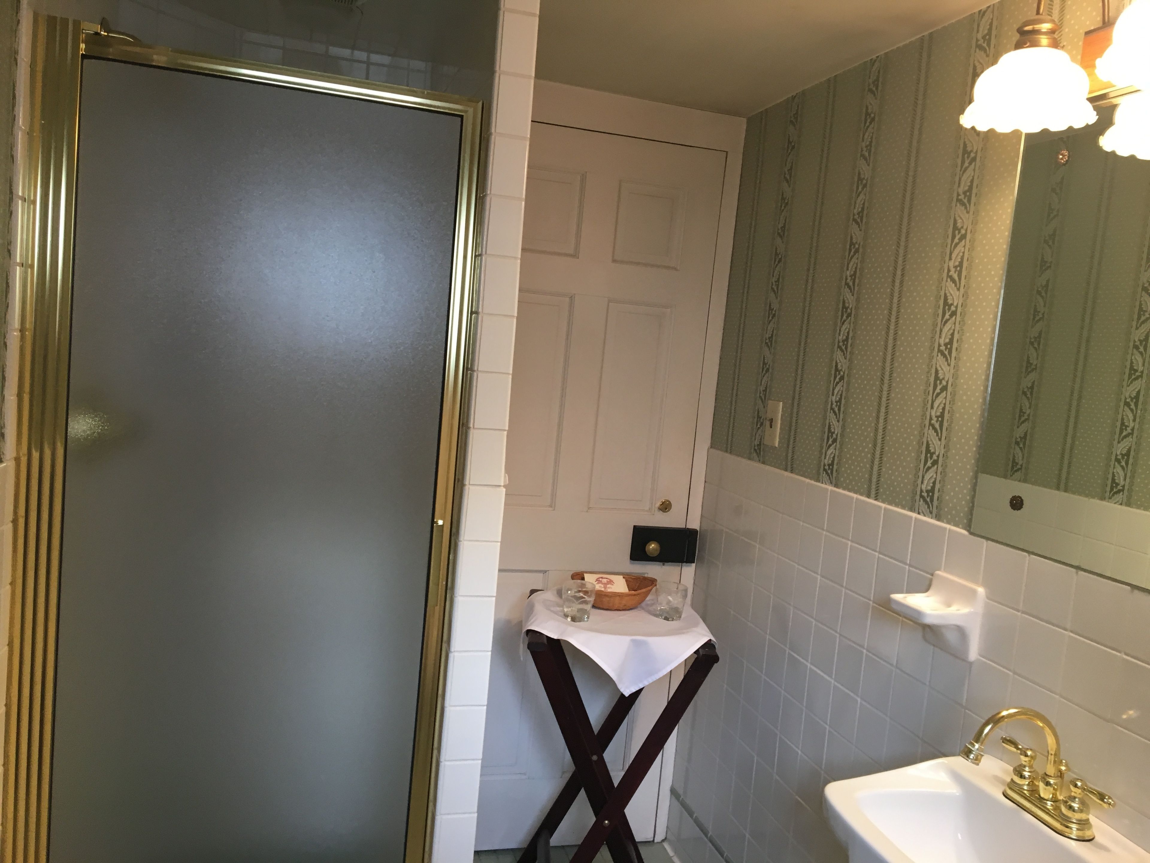Een badkamer met een witte wastafel, een gouden kraan en een spiegel erboven. Er staat een klein tafeltje met een mand en glazen, en een deur met een gouden omlijsting.