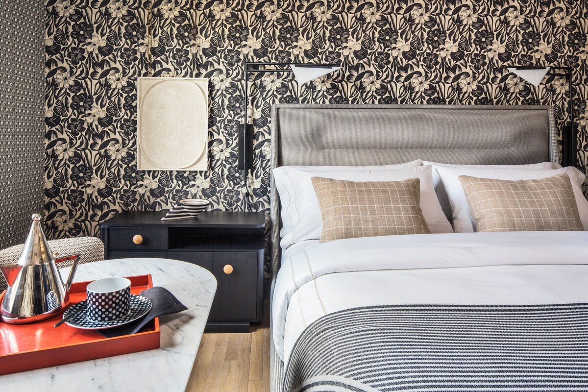 A neatly arranged bedroom with a large bed, a bedside table with a tray, a nightstand, and a wall with floral wallpaper.