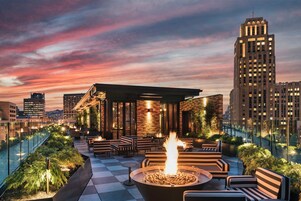 A rooftop terrace with a fire pit, seating, and a view of the city skyline at dusk.