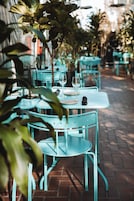 A row of outdoor tables with chairs, each set with a wine glass and a plate, surrounded by potted plants.