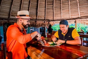 Dos personas cenando en una mesa de madera bajo un techo de paja.