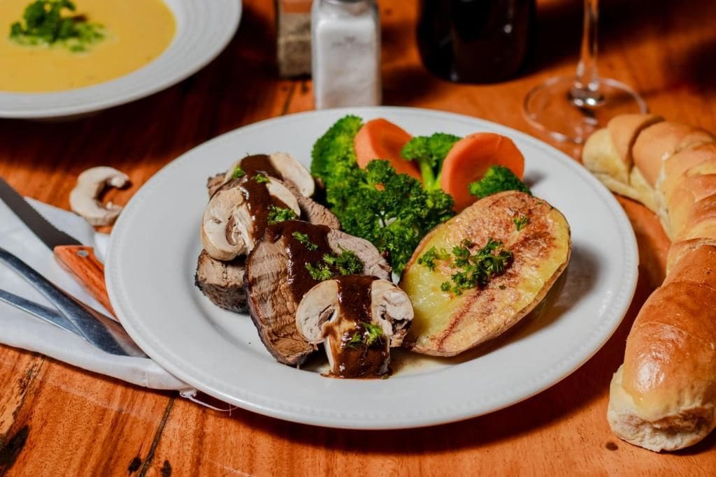 A plate with sliced meat, mushrooms, broccoli, and a roasted potato, accompanied by a bread roll and a glass of wine on a wooden table.