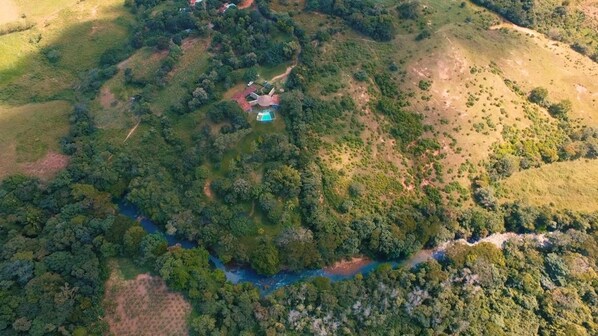 An aerial view of a property with a swimming pool surrounded by dense forest, a small building, and a winding river.