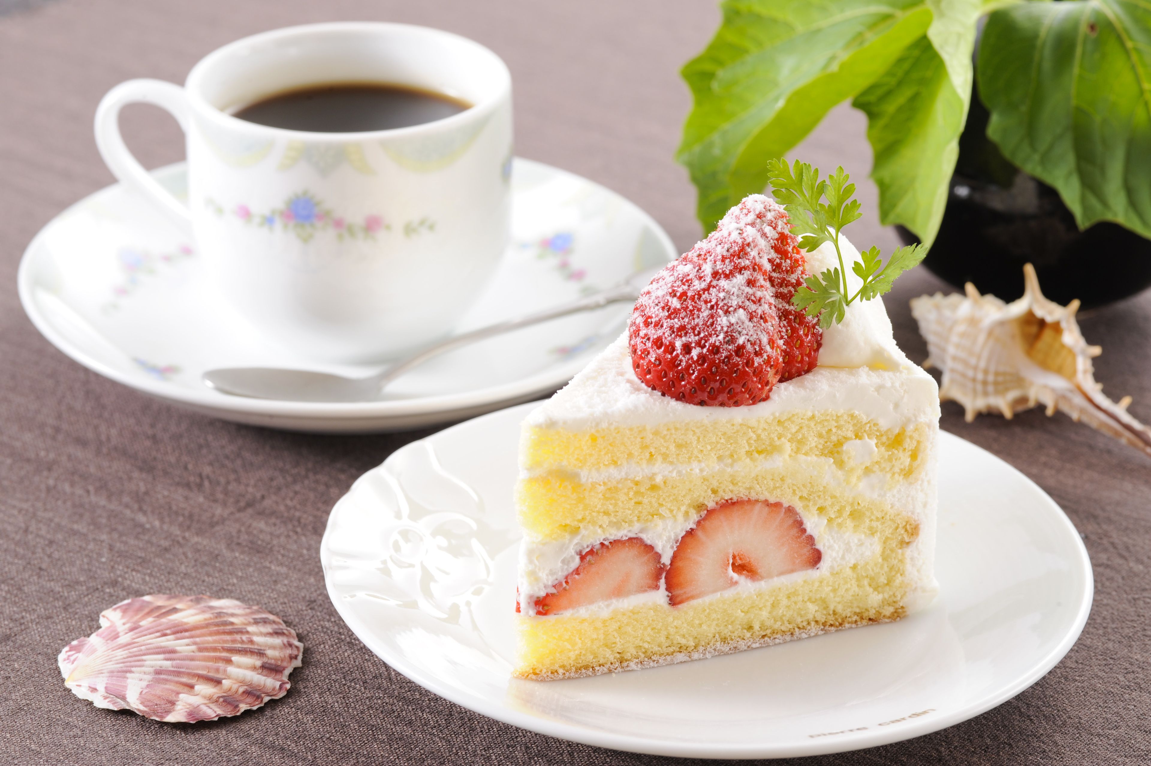 A slice of strawberry cake with cream frosting and a strawberry on top, served on a white plate with a cup of coffee in the background.