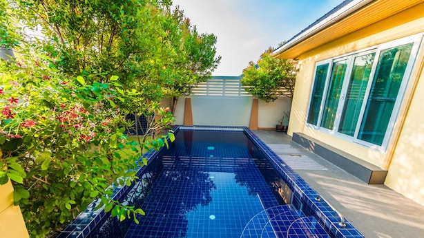 A rectangular swimming pool with blue tiles, surrounded by greenery and a building with large windows. - Platinum 3 bedrooms villa pool garden (Rawai)