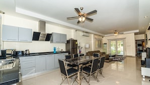 A modern kitchen with grey cabinets, a black countertop, and a dining area with a glass table and black chairs. - Platinum 3 bedrooms villa pool garden (Rawai)