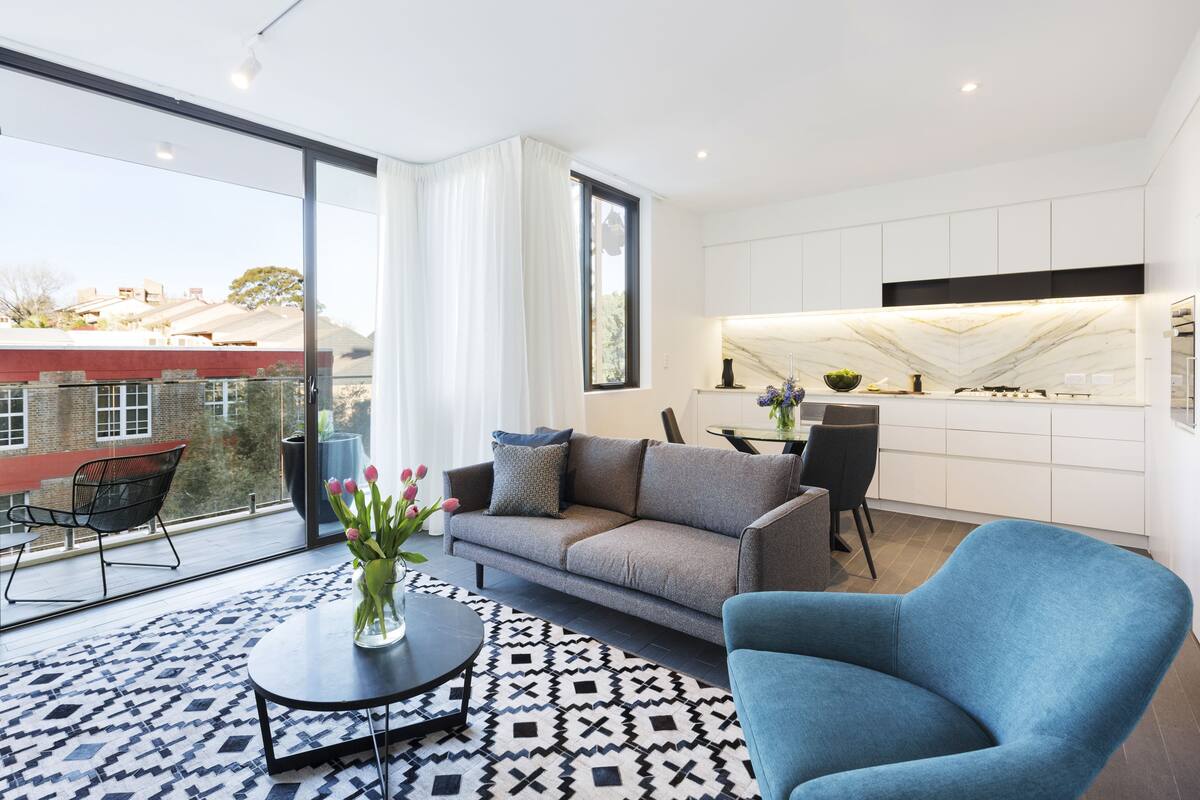 A modern living room with a grey sofa, a blue armchair, and a patterned rug. There is a dining area with a table and chairs, and a kitchen with white cabinets and a marble backsplash.