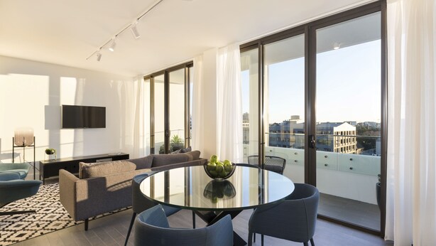 A modern living room with a round glass table, grey sofas, and a large window offering a city view. - Botanik Apartment Hotel (Surry Hills)