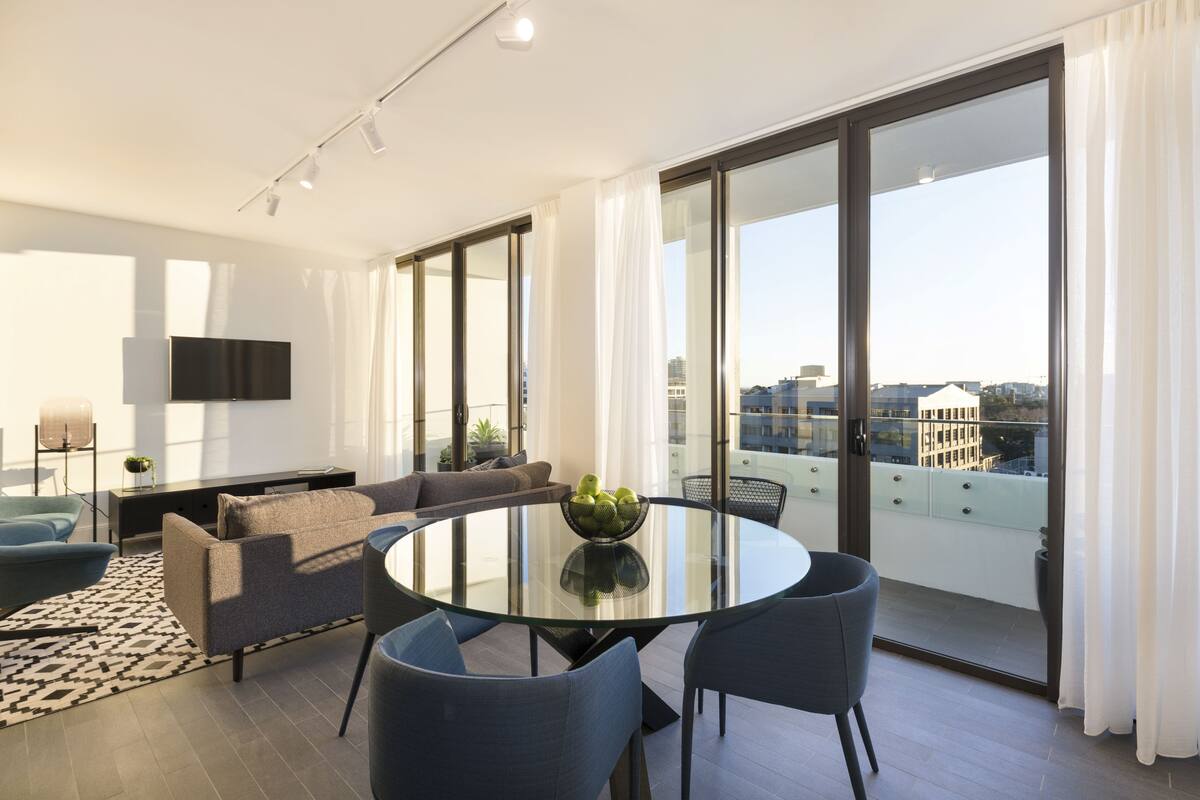 A modern living room with a round glass table, grey sofas, and a large window offering a city view.