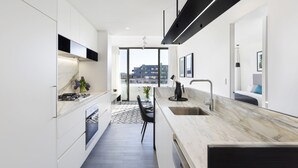 A modern kitchen with white cabinetry, a marble countertop, and a view of the city through large windows. - Botanik Apartment Hotel (Surry Hills)