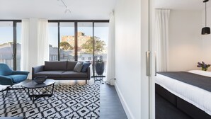 A modern hotel room with a large window, a sofa, a chair, a bedside table, and a bed. - Botanik Apartment Hotel (Surry Hills)
