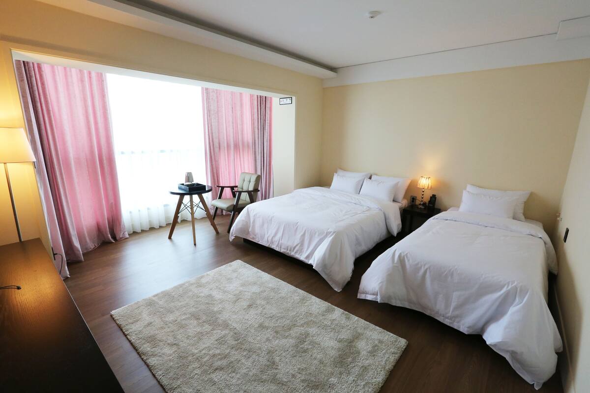 A hotel room with two beds, a small table, a chair, and a view of the outdoors through large windows with pink curtains.