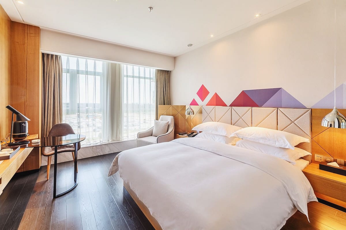 Une chambre d’hôtel avec un grand lit, un bureau avec une chaise, une lampe de bureau et une vue sur la ville grâce à de grandes fenêtres.