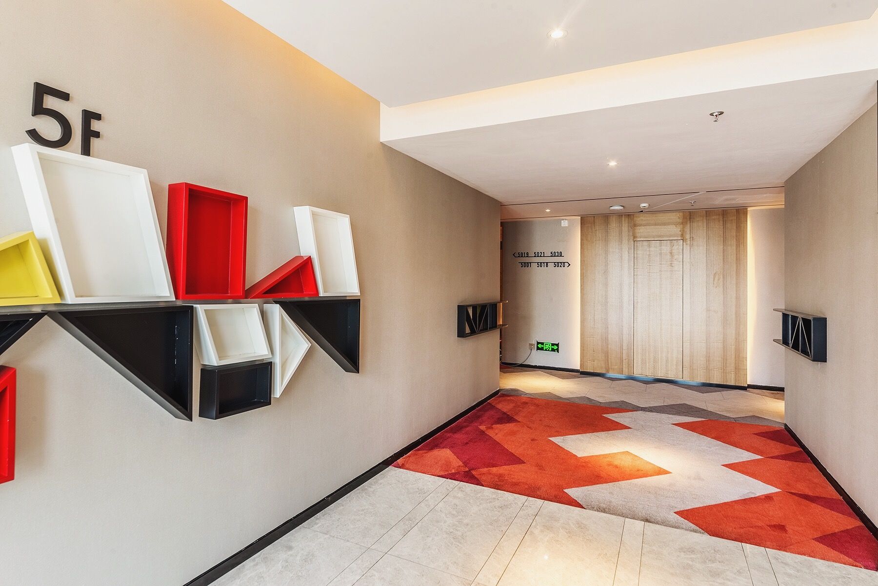 A hallway with a geometric patterned carpet, a wall-mounted shelf with variously colored objects, and a door with directional signage.