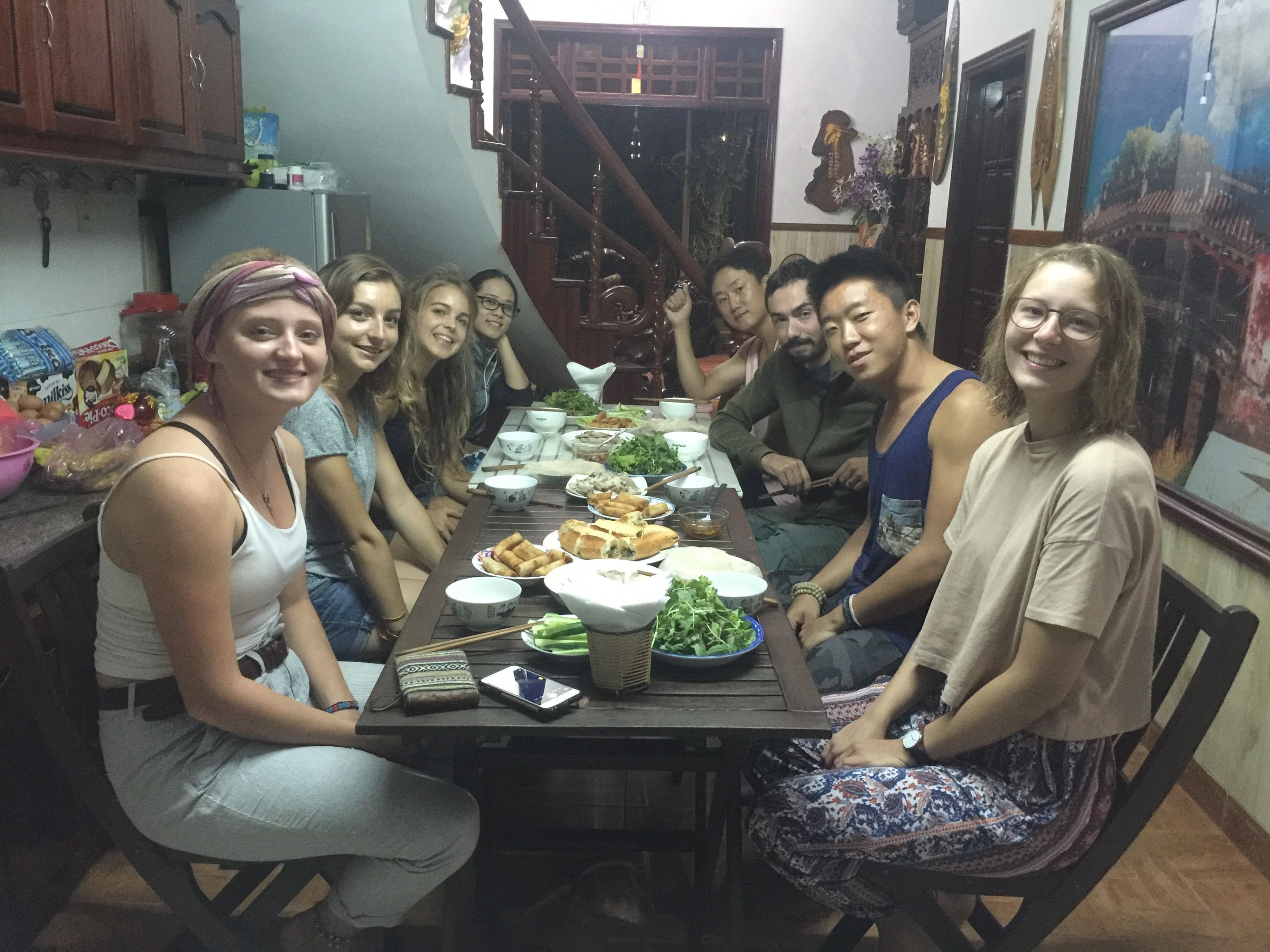 A group of people seated around a table with various dishes, in a room with wooden furniture and a staircase.