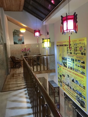 A hotel lobby with a staircase, wooden railing, and hanging lanterns. There is a poster with tourist information on the wall.