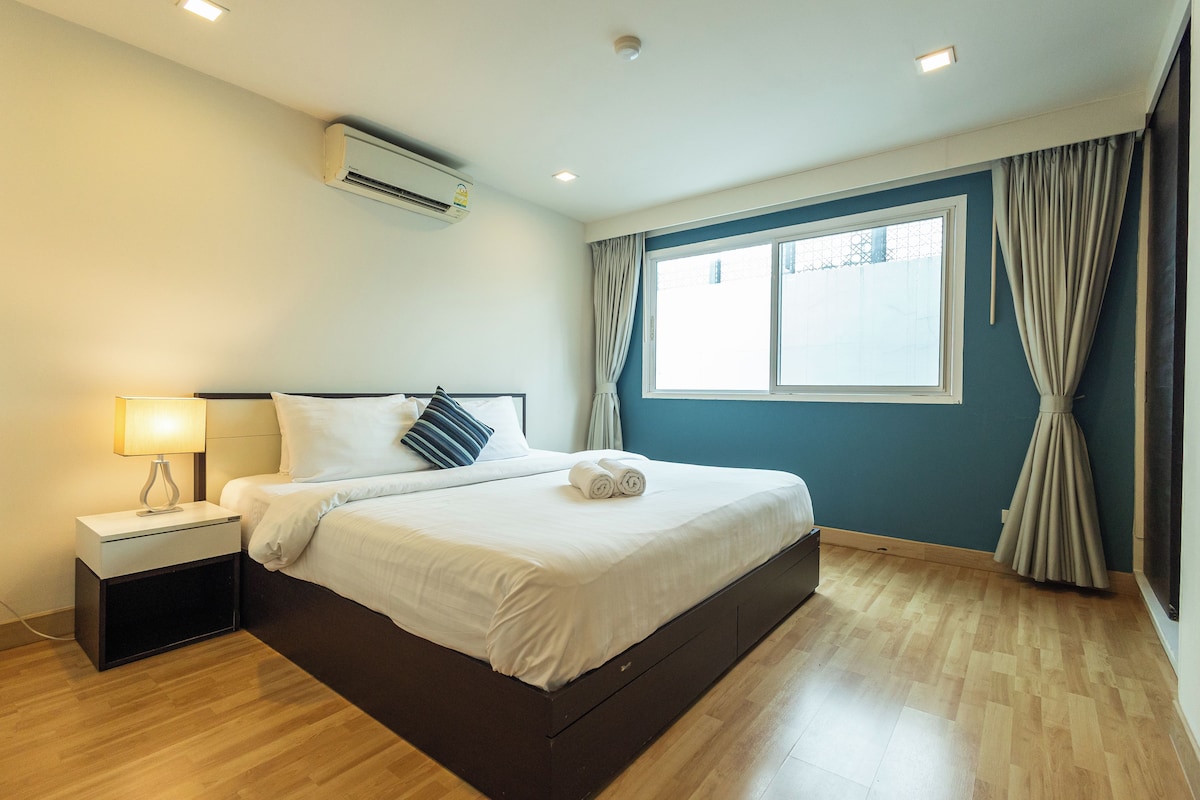 A hotel room with a bed, bedside table, lamp, air conditioning unit, window with curtains, and wooden flooring.