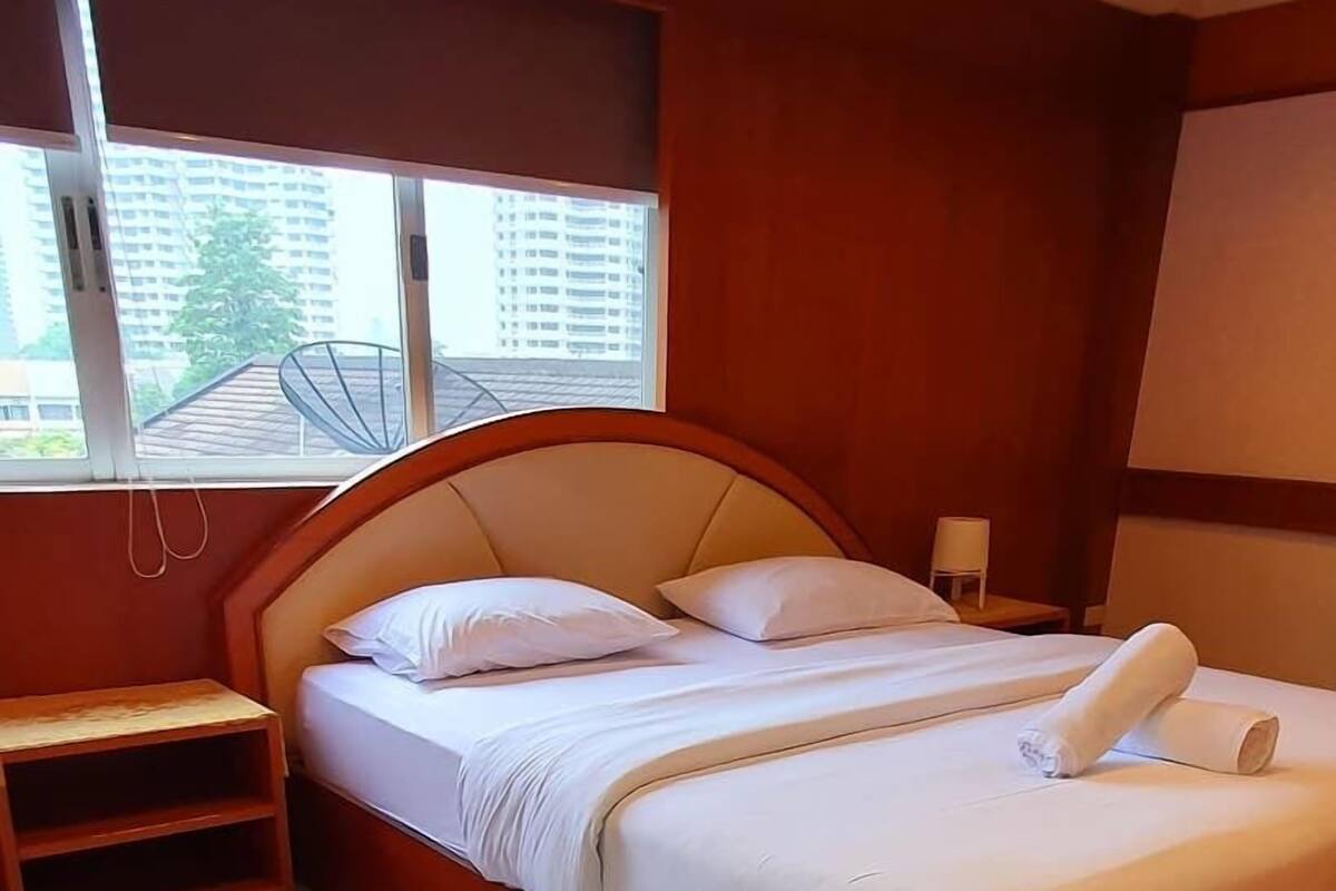 Una habitación de hotel con una cama grande, una mesita de noche, un ventanal con vistas a edificios y un piso de baldosas.