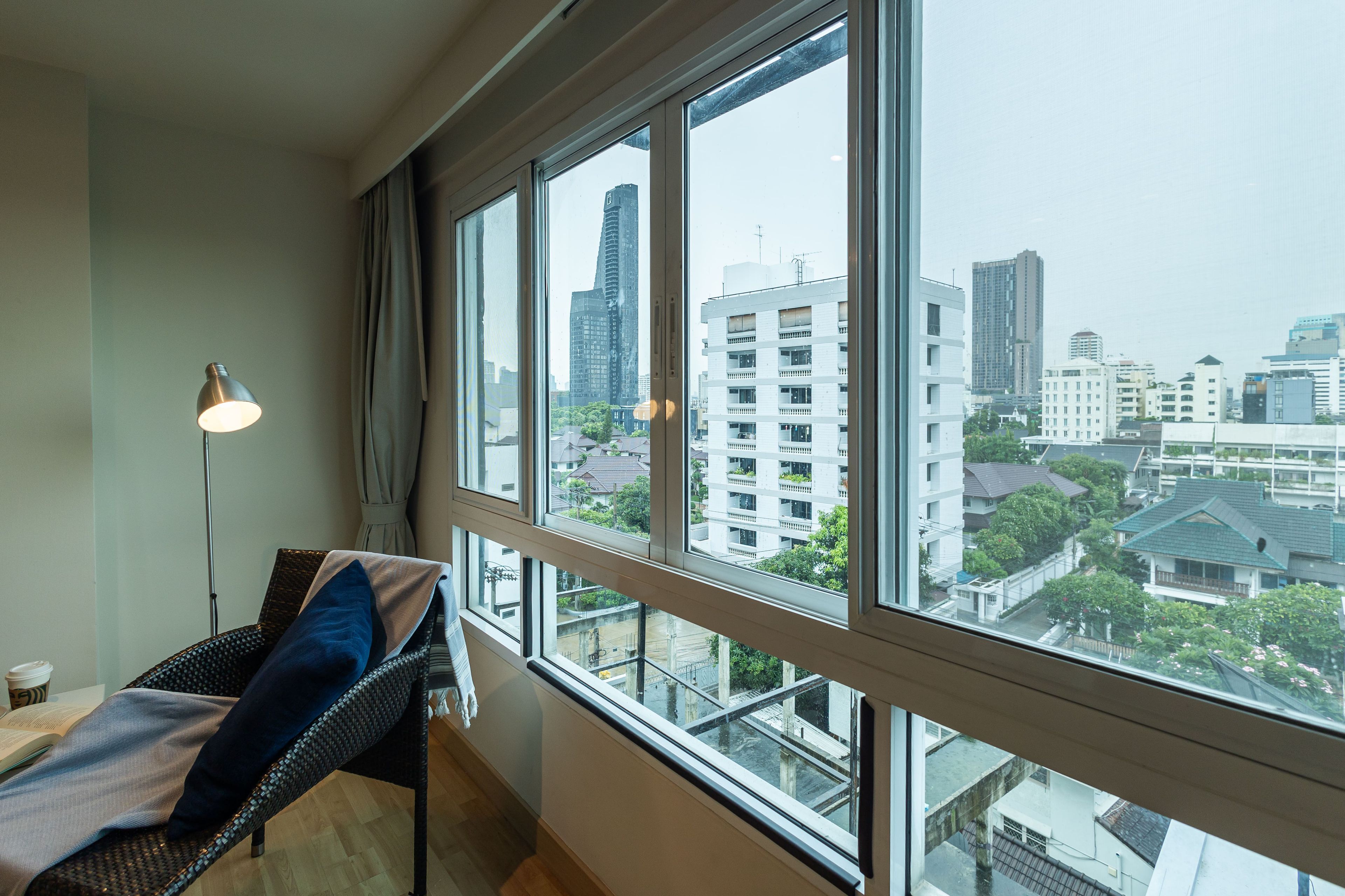 A room with a large window, a wicker chair, a lamp, and a view of city buildings.