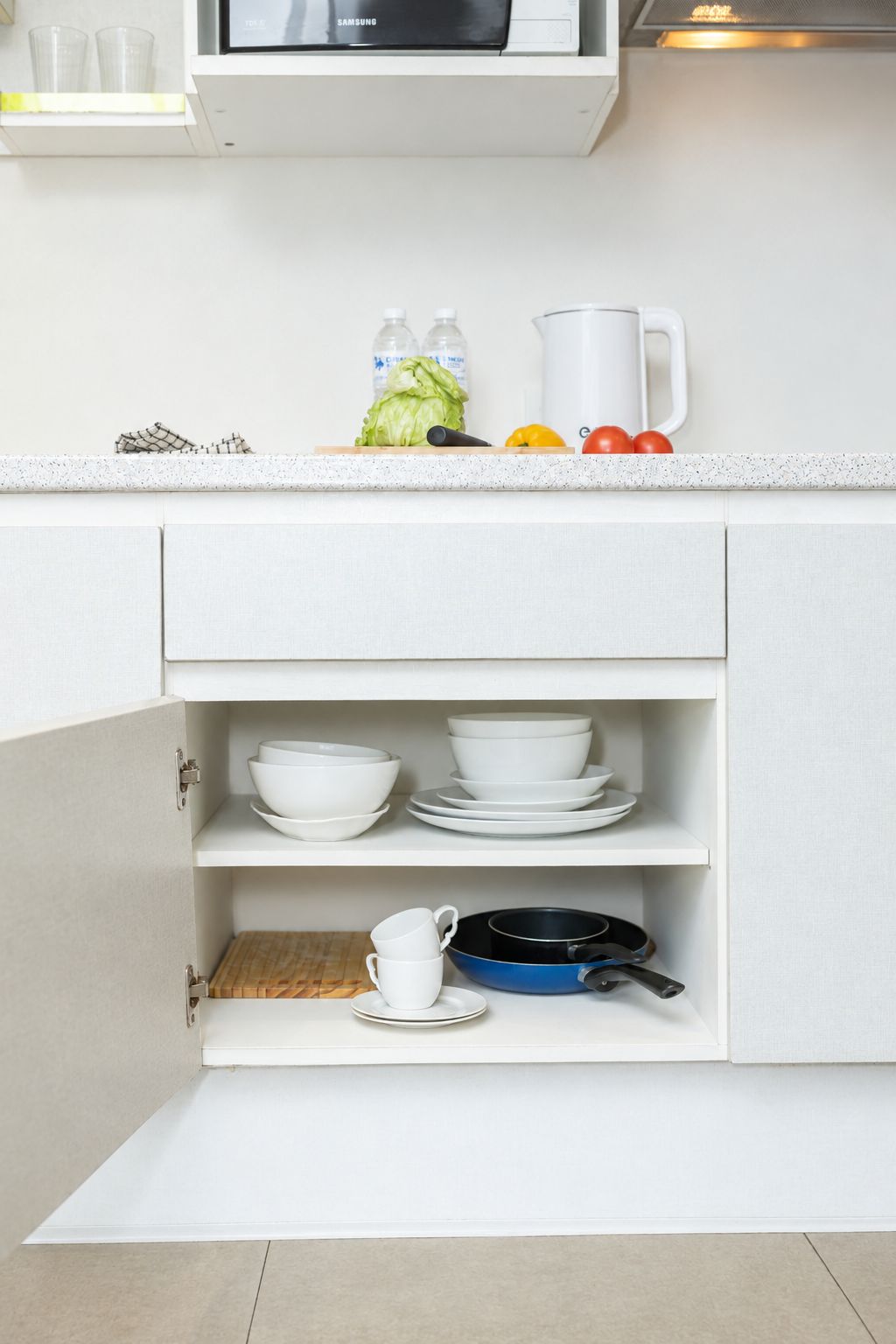 A kitchen with white cabinets, a microwave, and a countertop with bottles, a kettle, and vegetables.