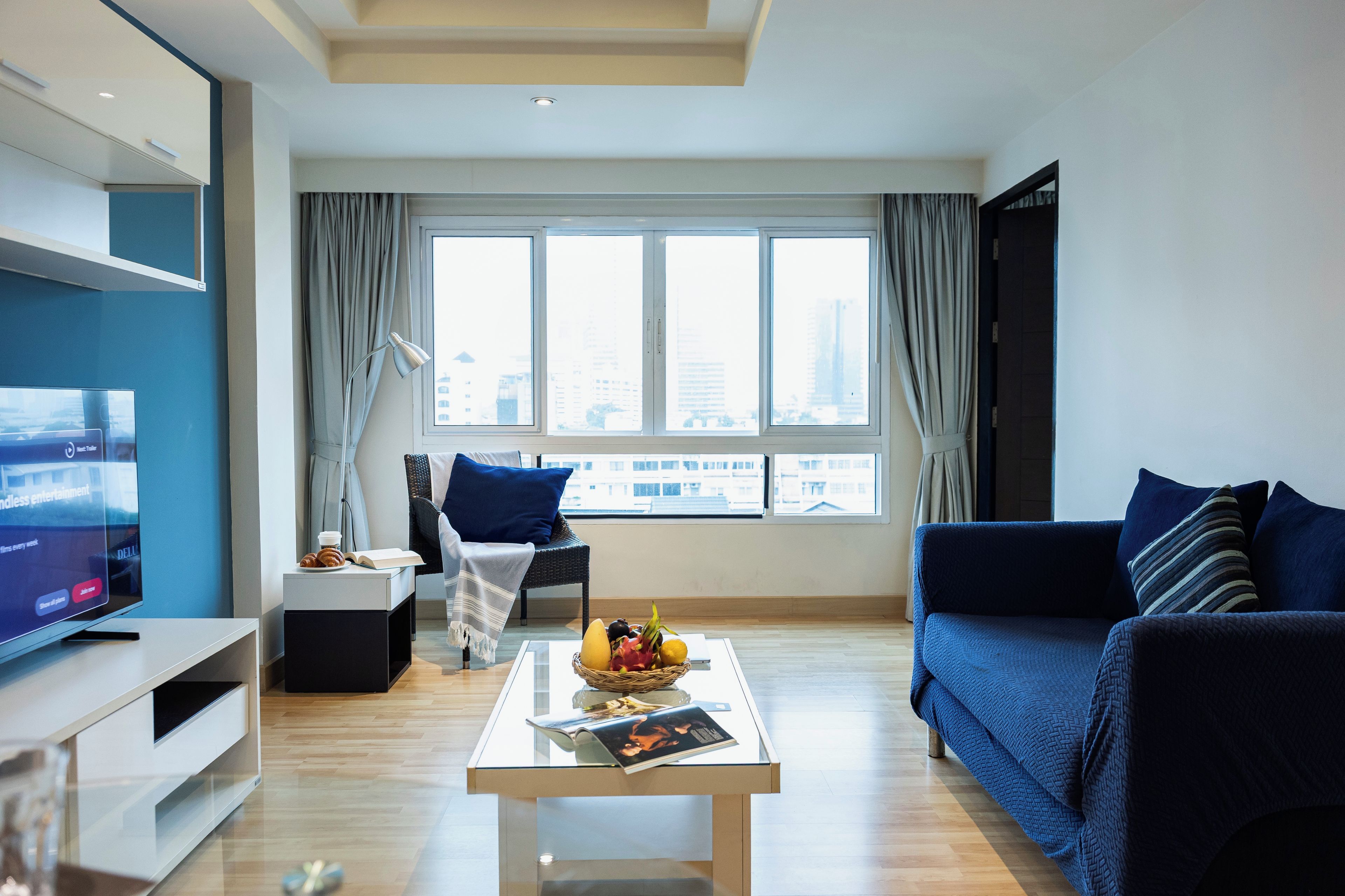 A modern living room with a television, a coffee table with a fruit basket, a sofa, and a view of the city through large windows.