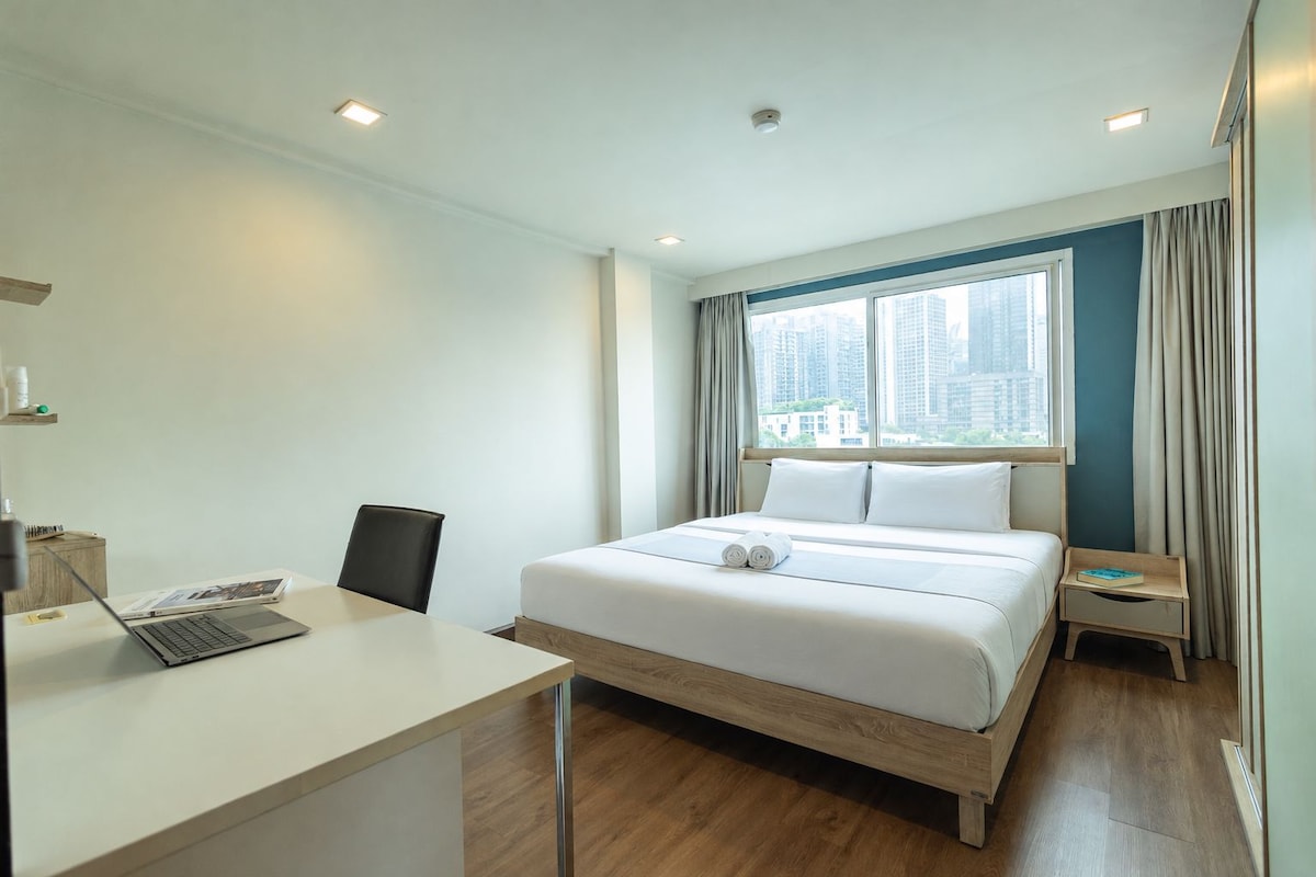 A hotel room with a bed, a desk with a laptop, a chair, and a nightstand with a book.