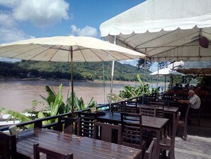 Un restaurant situé au bord de la rivière, doté d’une terrasse, de grands parasols et offrant une vue sur la rivière et les collines boisées.