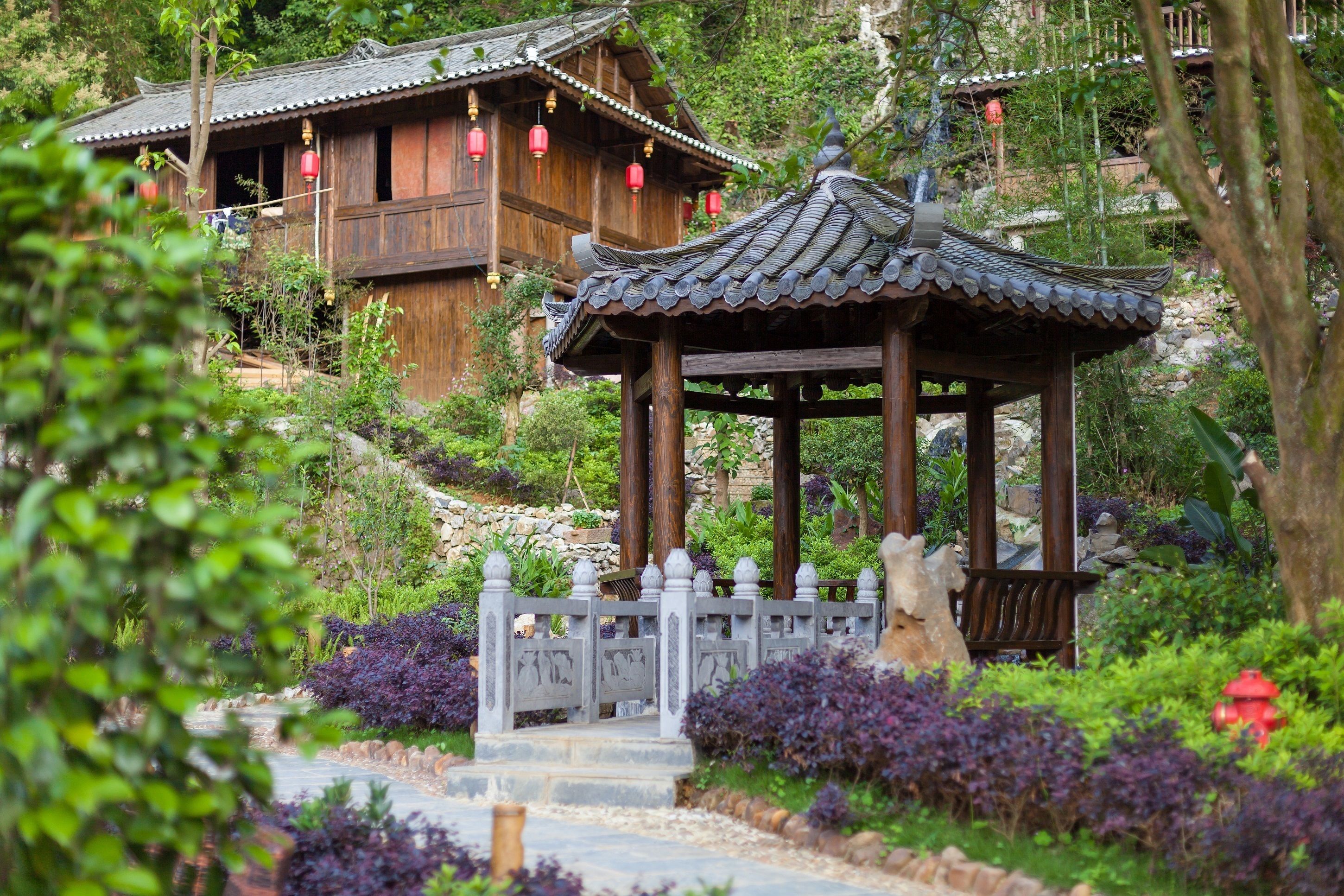A traditional wooden building with a tiled roof and red lanterns, surrounded by lush greenery and a stone pathway.