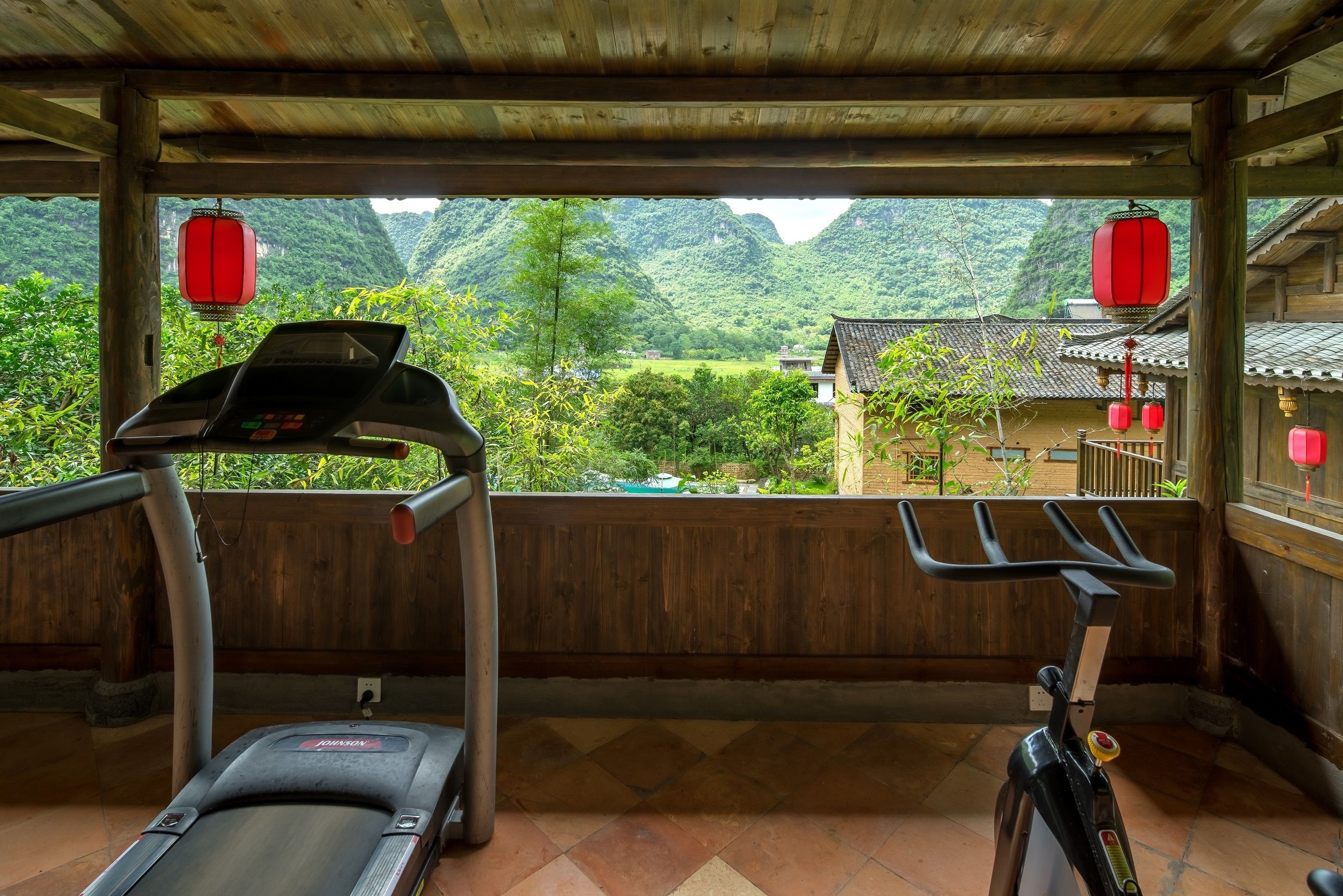 Overdekte trainingsruimte met loopband en hometrainer, met uitzicht op een traditioneel dorp met rode lantaarns en terrassen.