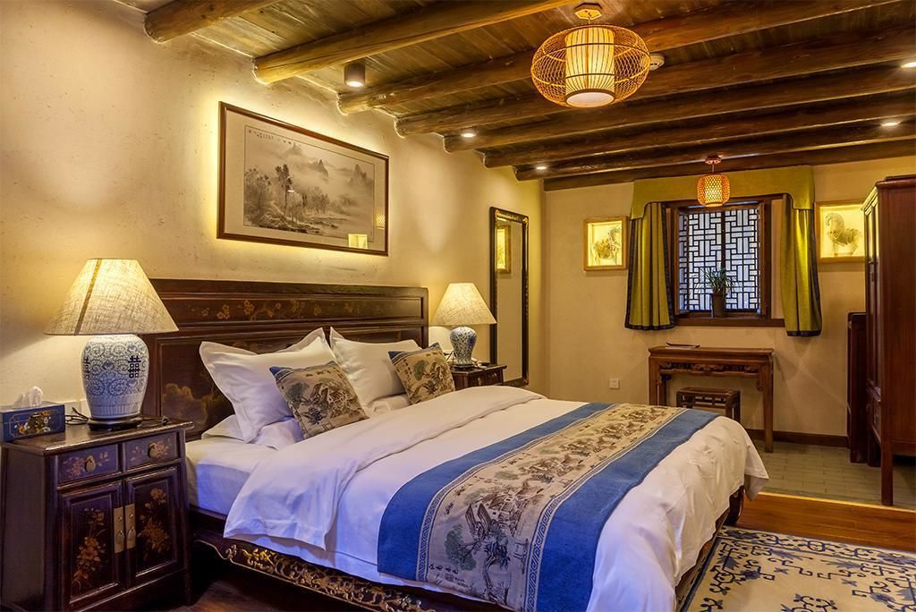 Un dormitorio tradicional con cama de madera, dos mesitas de noche con lámparas, un cuadro grande enmarcado en la pared y una ventana con celosía decorativa.