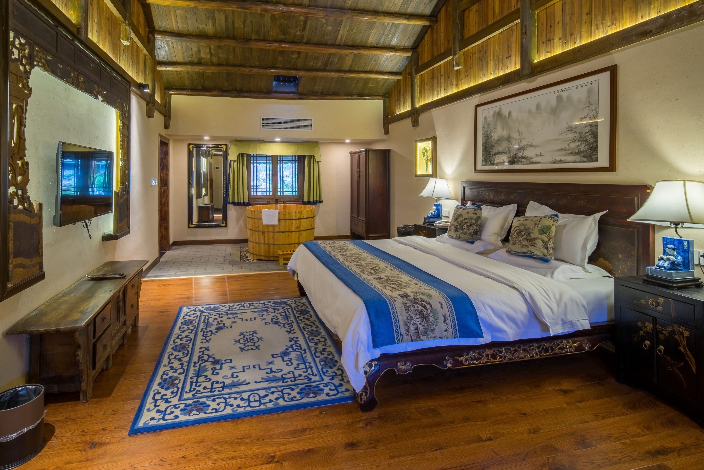 A traditional room with a large bed, wooden flooring, a decorative rug, and a painting on the wall.