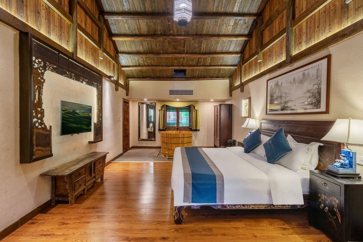 A spacious room with a large bed, wooden flooring, and a high ceiling with exposed beams.