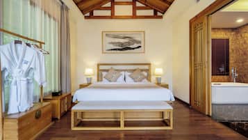 A bedroom with a wooden bed, bedside lamps, a wooden bench, a robe rack, and a bathroom with a bathtub.