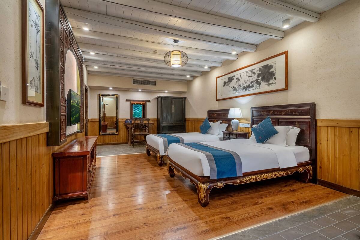 A hotel room with two beds, wooden flooring, a mirror, a desk, and framed artwork on the walls.