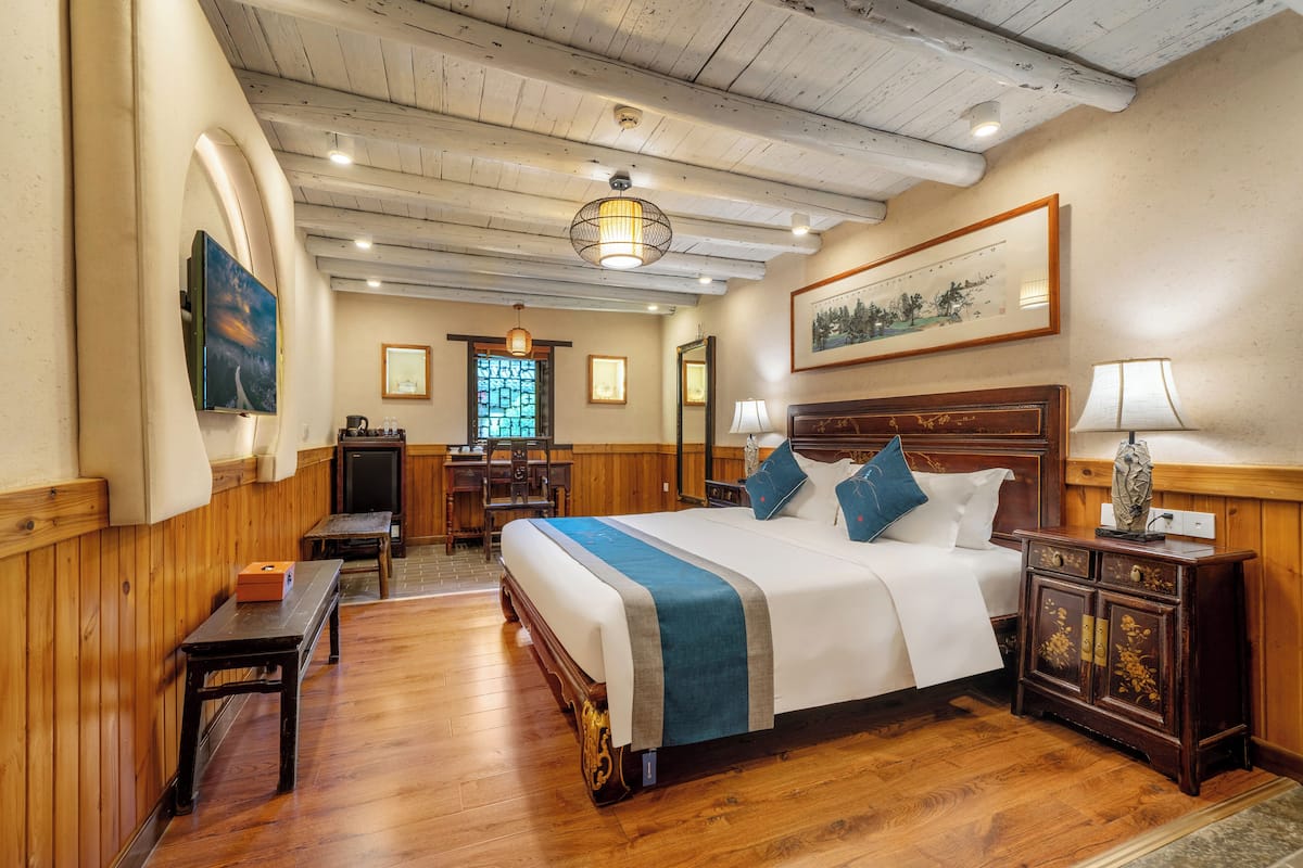 A hotel room with a wooden floor, a large bed with a blue and white bedspread, a wooden nightstand with a lamp, a desk with a chair, and a window with a view of trees.