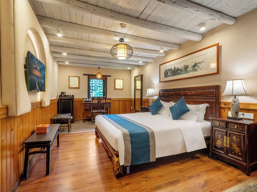 A hotel room with a wooden floor, a large bed with a blue and white bedspread, a wooden nightstand with a lamp, a desk with a chair, and a window with a view of trees.