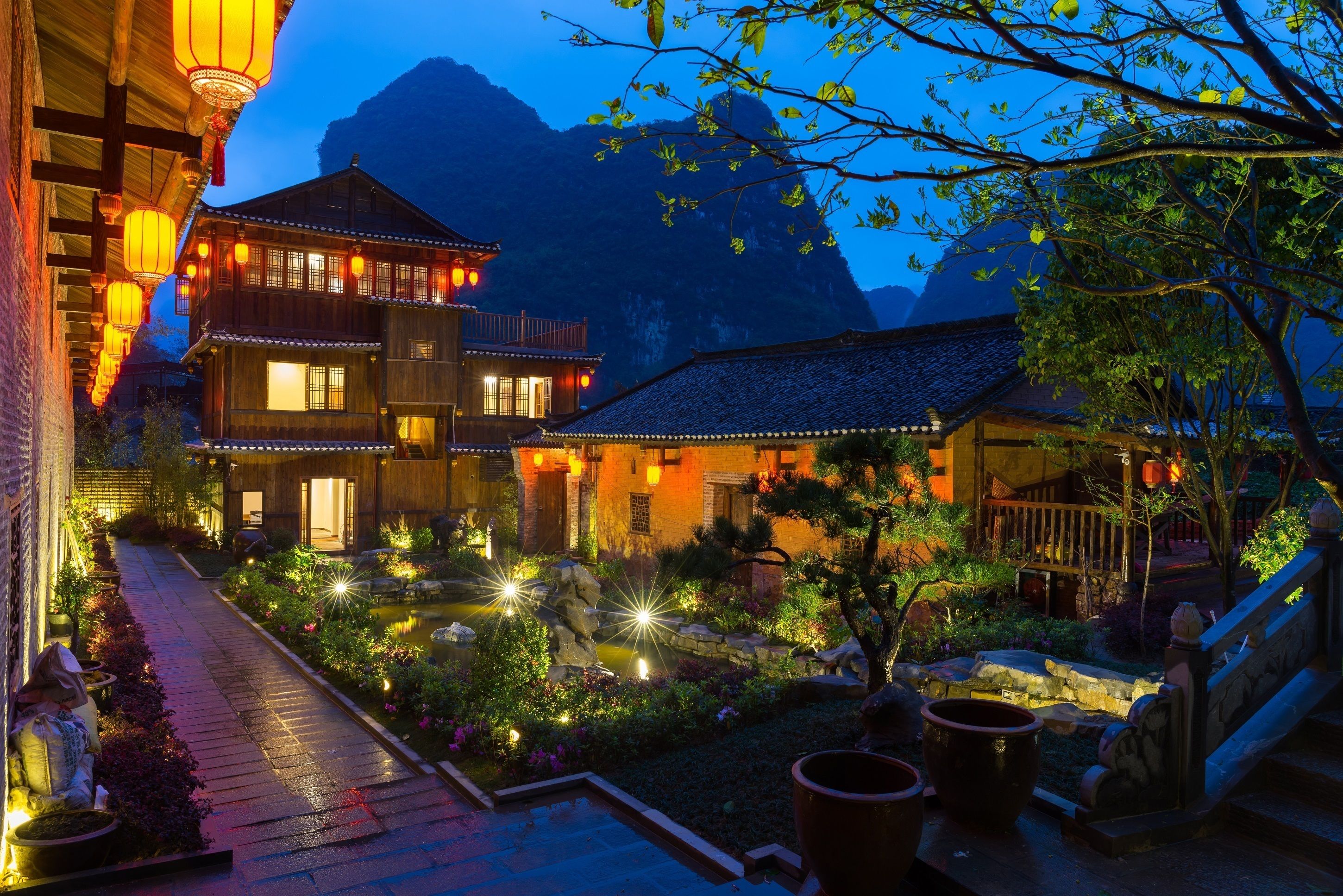 A traditional wooden house with a garden pathway, a pond, and a mountain backdrop.
